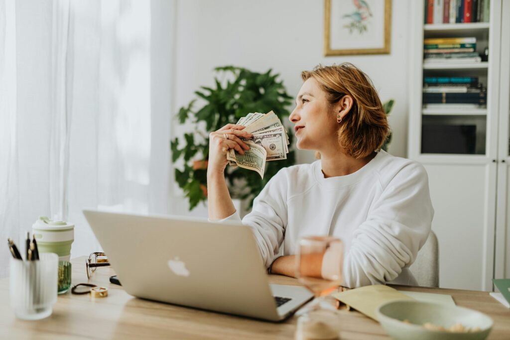 pexels-photo-5899215-5899215 Woman sitting at desk holding cash while working on a laptop in a cozy home office setting.
