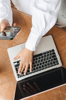 pexels-photo-7382449-7382449 Hands typing on a laptop while holding a smartphone, showing a multitasking work environment.